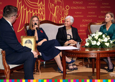 siedzący Mężczyzna i trzy kobiety, jedna z nich mówi przez mikrofon