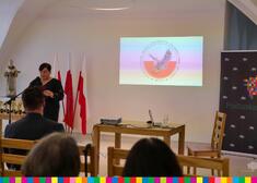 Kobieta przemawia, za nią statuetki, flagi biało czerwone oraz wyświetlany slajd multimedialny