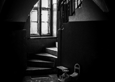 Czarno biała fotografia schodów prowadzonych w góre, przy których jest oświetlone okno