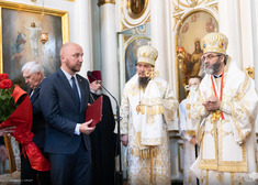 marszałek województwa stoi przy mikrofonie, przed nim dwóch duchownych