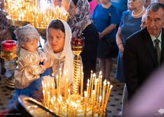 Kobieta w chustce trzyma dziecko na rękach, przednimi i w tle stoją świeczniki z cienkimi zapalonymi świeczkami