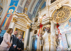Duchowny trzyma w rękach świeczniki z zapalonymi świecami, obok stoją samorządowcy