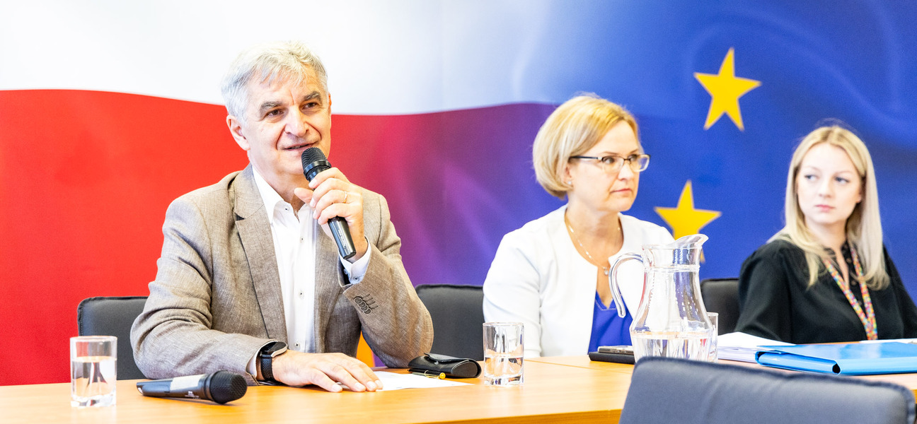 Bogdan Dyjuk siedzi obok kobiety na tle flag Polski i Unii.