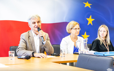 Bogdan Dyjuk siedzi obok kobiety na tle flag Polski i Unii.