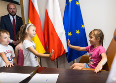 Dzieci przybijają piątkę na tle flag unii, polski i województwa.