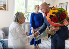 Marszałek wita się ze starszą Panią przekazując jej bukiet kwiatów. W tle stoi wicemarszałek