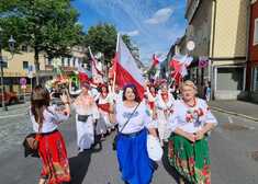grupa kobiet w strojach ludowych maszeruje ulicą; trzymają polskie flagi