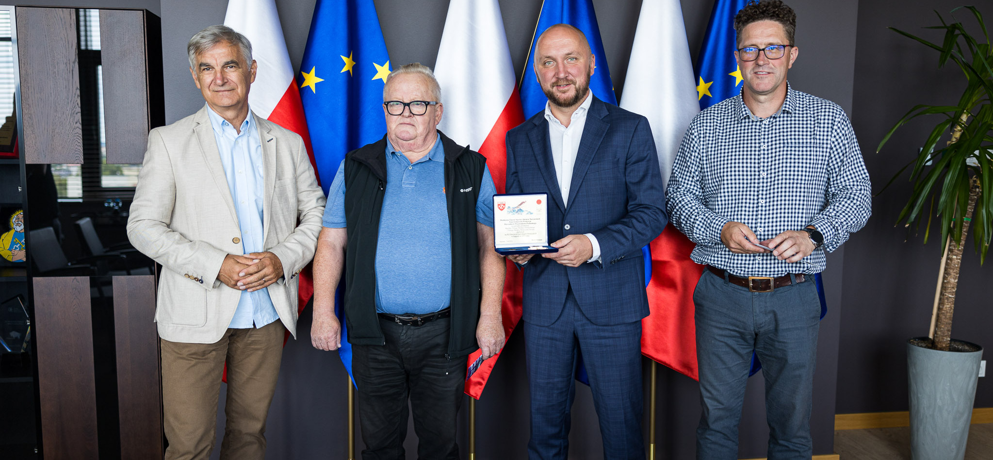 Dwóch przedstawicieli zarządu województwa oraz Wojciech Fortuna i prezes WOSiRu stoją na tle flag