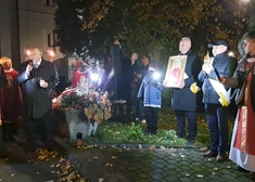 Kilkanaście osób stoi, oddając hołd księdzu Jerzemu Popiełuszko. jedna osoba trzyma portret księdza. jedna osoba przemawia.