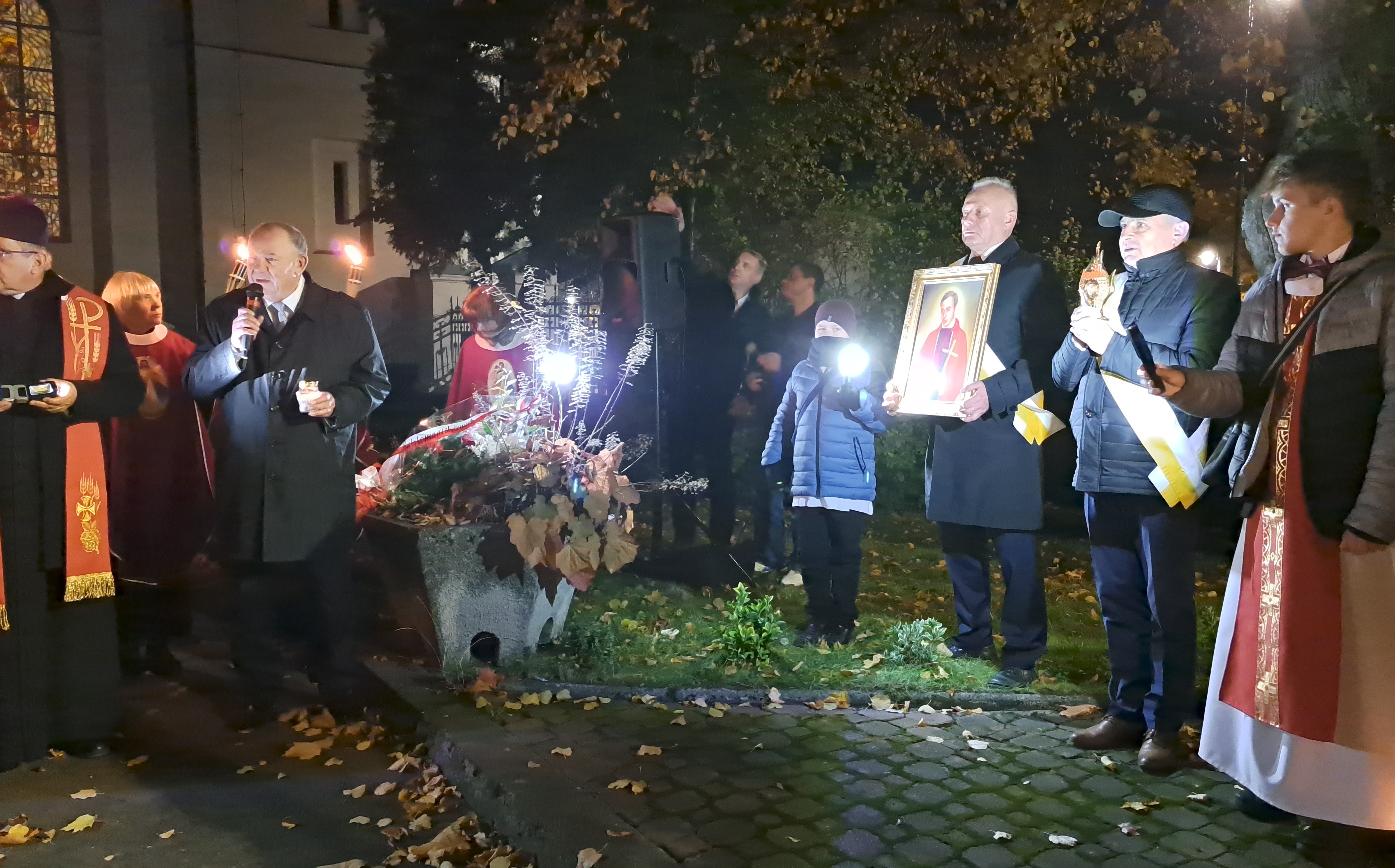 Kilkanaście osób oddaje hołd księdzu Jerzemu Popieuszko. Jedna osoba trzyma w ręku portret księdza. Jedna osoba przemawia.