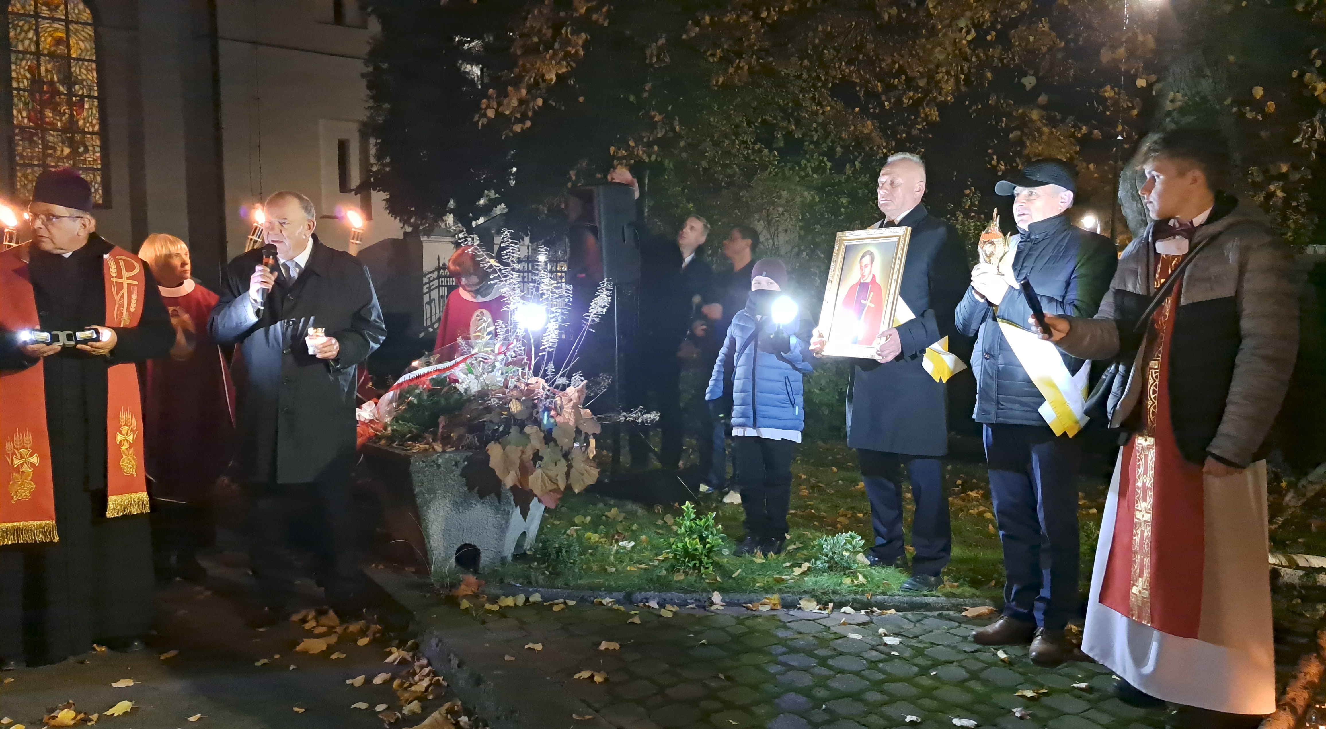 Kilkanaście osób oddaje hołd księdzu Jerzemu Popieuszko. Jedna osoba trzyma w ręku portret księdza. Jedna osoba przemawia.