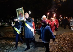 Kolejne osoby idą w pochodzie ze zniaczami. Mężczyna trzyma wysoko uniesiony portret księdza Jerzego Popiełuszko.