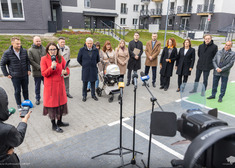 Jedna osoba przemawia, kilkanaście słucha, w tle widok bloków mieszkaniowych