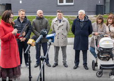 Ta sam osoba przemawia, kilkanaście słucha, w tle widok bloków mieszkaniowych