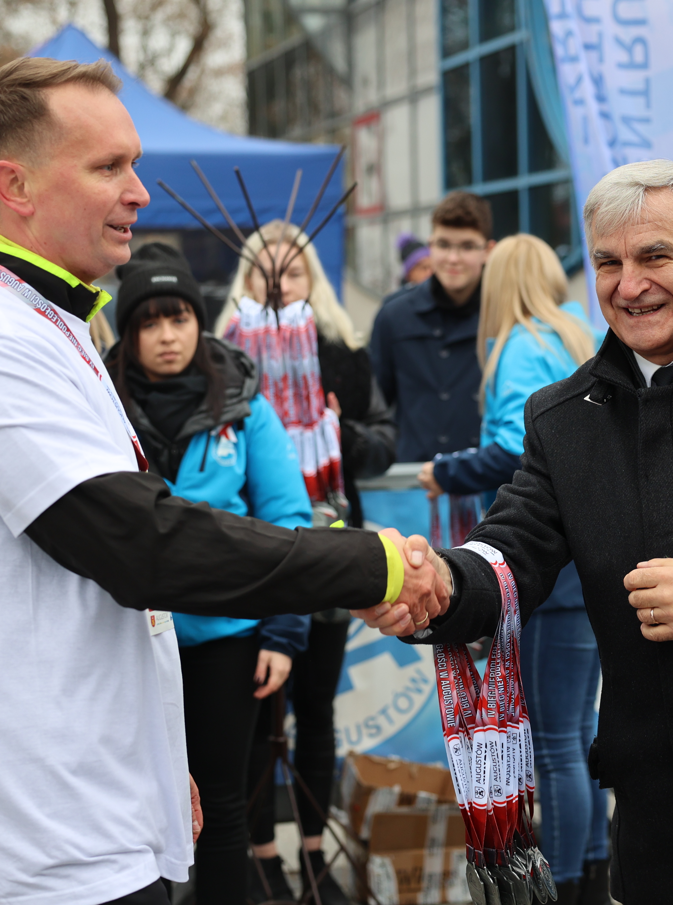 Dwaj mężczyźni podają sobie ręce. Mężczyzna po prawej ubrany w białą sportową koszulkę, mężczyzna po lewej w ciemną kurtkę, z przodu ma przypięty biało-czerwony kotylion