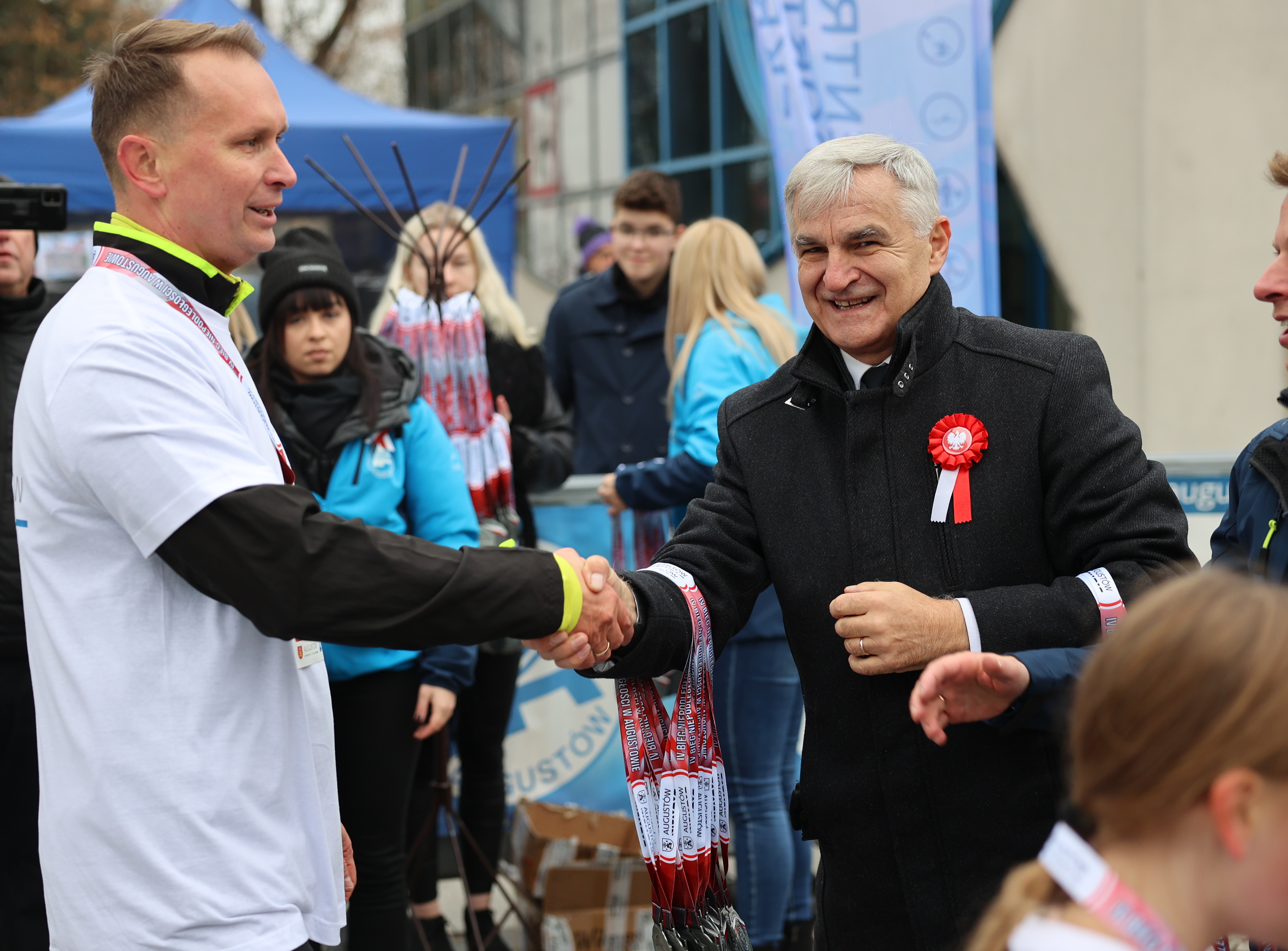 Dwaj mężczyźni podają sobie ręce. Mężczyzna po prawej ubrany w białą sportową koszulkę, mężczyzna po lewej w ciemną kurtkę, z przodu ma przypięty biało-czerwony kotylion