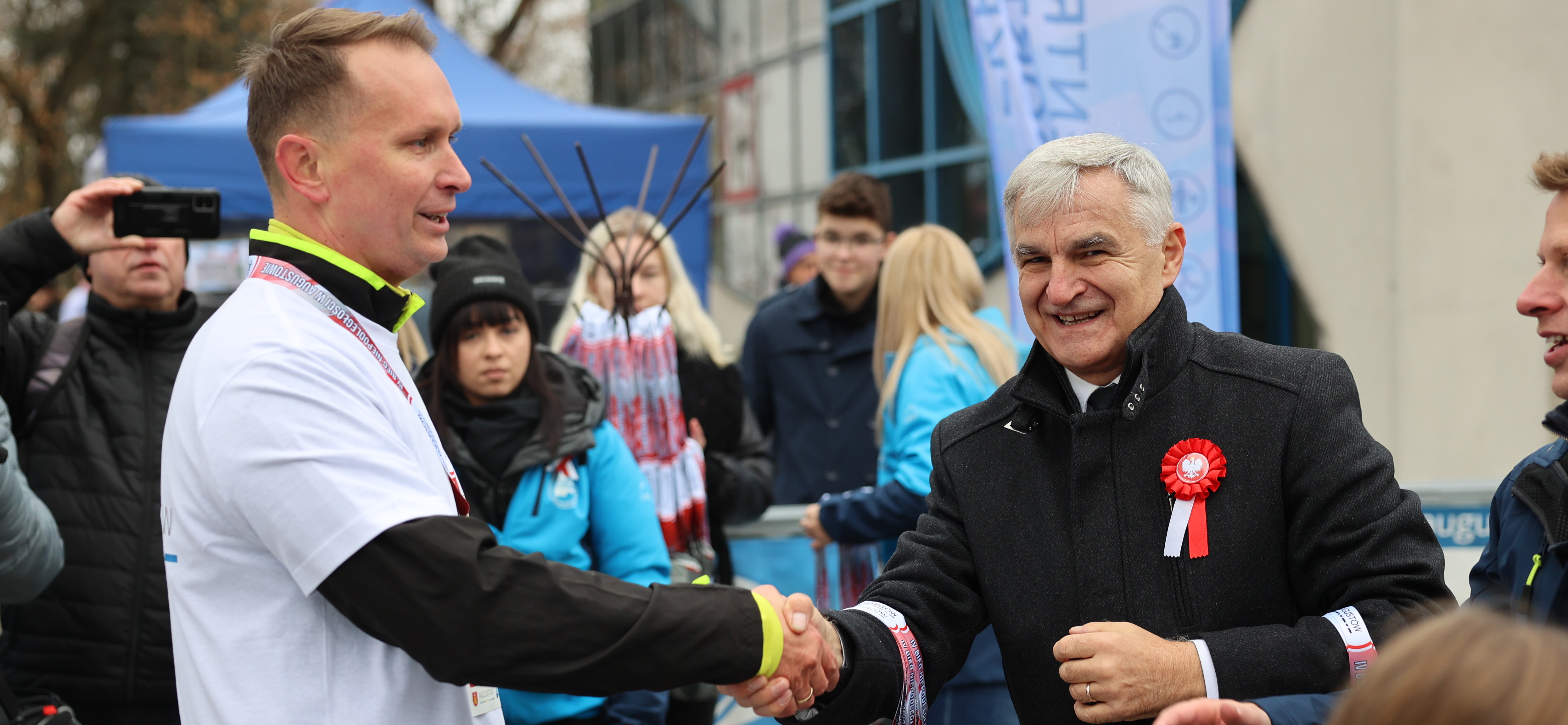 Dwaj mężczyźni podają sobie ręce. Mężczyzna po prawej ubrany w białą sportową koszulkę, mężczyzna po lewej w ciemną kurtkę, z przodu ma przypięty biało-czerwony kotylion