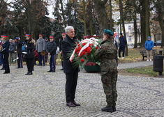 Mężczyzna w ciemnym ubraniu przekazuje wieniec z czerwonych kwiatów z białą wstążką kobiecie w mundurze