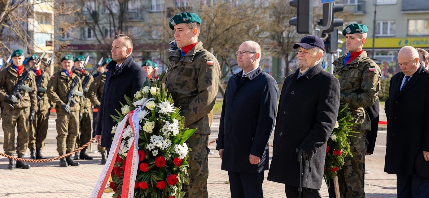 Trzech elegancko ubranych mężczyzn stoją w otoczeniu żołnierzy. Jeden z żołnierzy przytrzymuje wieniec z kwiatów