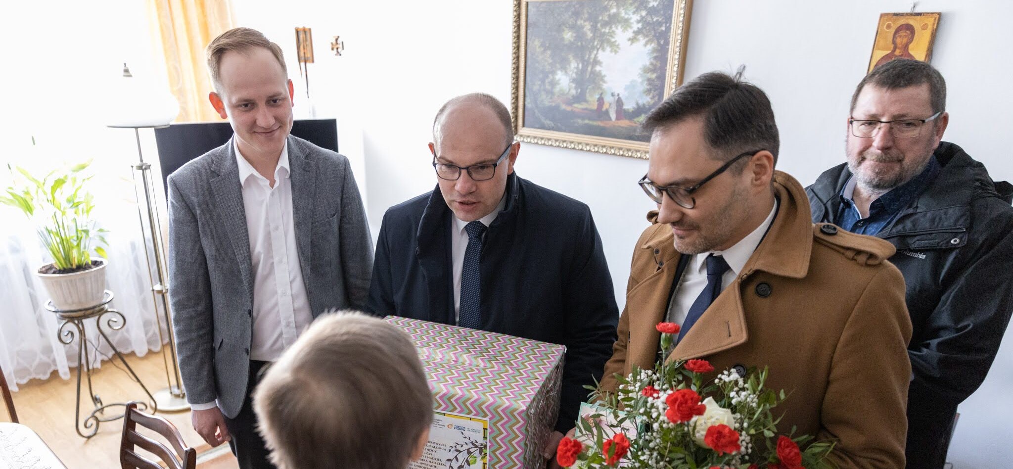 Czterej mężczyźni z prezentami i kwiatami stoją przed starszą kobieta.