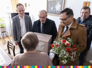 Czterej mężczyźni z prezentami i kwiatami stoją przed starszą kobieta.