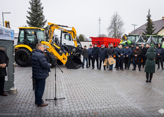 Mężczyzna mówi do mikrofonu w tle koparko-ładowarka i ludzie