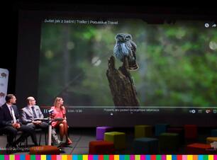 Trzy osoby na scenie, w tle ekran z sową siedzącą na pniu drzewa.