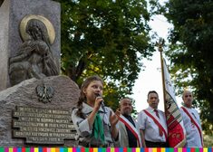 Przy Pomniku Katyńskim stoi dziewczynka w mundurze harcerskim, trzyma mikrofon przy ustach. Po prawej w tle stoi trzech mężczyzn z przewiązanymi szarfami biało-czerwonymi, jeden z nich trzyma sztandar.