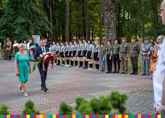 Kobieta w niebieskiej sukience i mężczyzna w garniturze idą w kierunku pomnika. Mężczyzna w rękach trzyma wiązankę kwiaty w kolorach biało-czerwonych. Po prawej w rzędzie stoją mężczyźni w mundurach, harcerze i pozostali uczestnicy wydarzenia.
