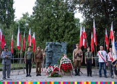 Pomnik Inki w Parku Planty. Przed pomnikiem złożone wiązanki kwiatów w barwach biało-czerwonych. Po obu stronach pomnika stoją mężczyźni w mundurach i harcerze. Po lewej stoi mężczyzna w mundurze przy mikrofonie, po lewej stoi trzech mężczyzn z przewiązanymi szarfami biało-czerwonymi, jeden z nich trzyma sztandar. Po obu stronach w tle stoją flagi biało-czerwone.