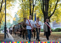 Trzech mężczyzn przewiązanych biało-czerwonymi wstążkami, jeden z nich trzyma biało-granatowy sztandar, za nimi w rzedach mężczyźni w mundurach i młodzież w mundurach harcerskich oraz pozostali uczestnicy wydarzenia. 