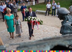 Kobieta w niebieskiej sukience trzyma za rękę harcerkę. Tuż obok nikt stoi mężczyzna trzymający wieniec