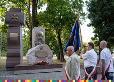 Przed Pomnikiem Katyńskim w Parku Konstytucji 3 Maja stoi trzech mężczyzn przewiązanych szarfami biało-czerwonymi. Mężczyzna stojący pośrodku trzyma w rękach granatowy sztandar.