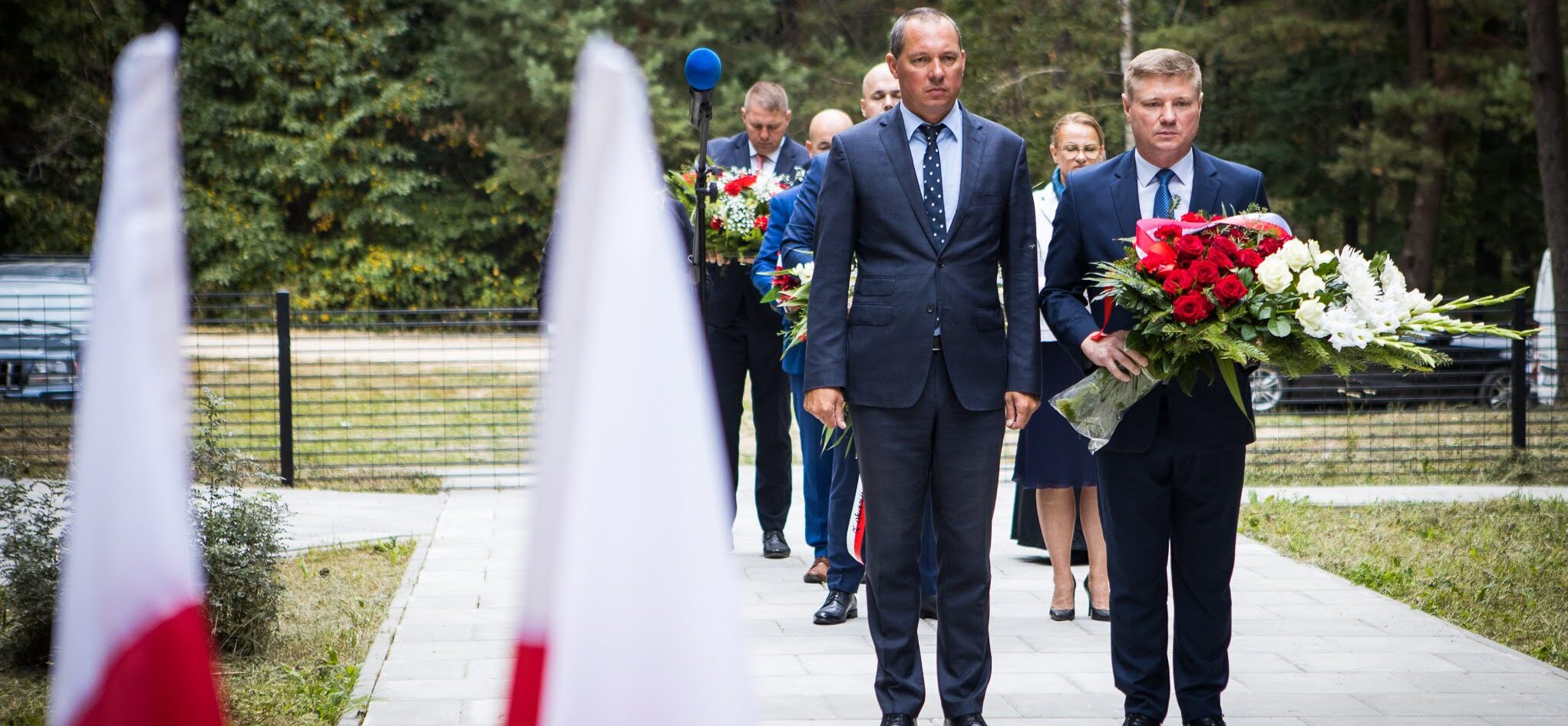 Kilka osób w odświętnych strojach niesie biało-czerwone kwiaty