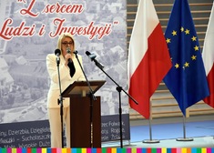 Kobieta w kremowym garniturze mówi do mikrofonu. Po prawej stronie flaga Polski i Unii Europejskiej