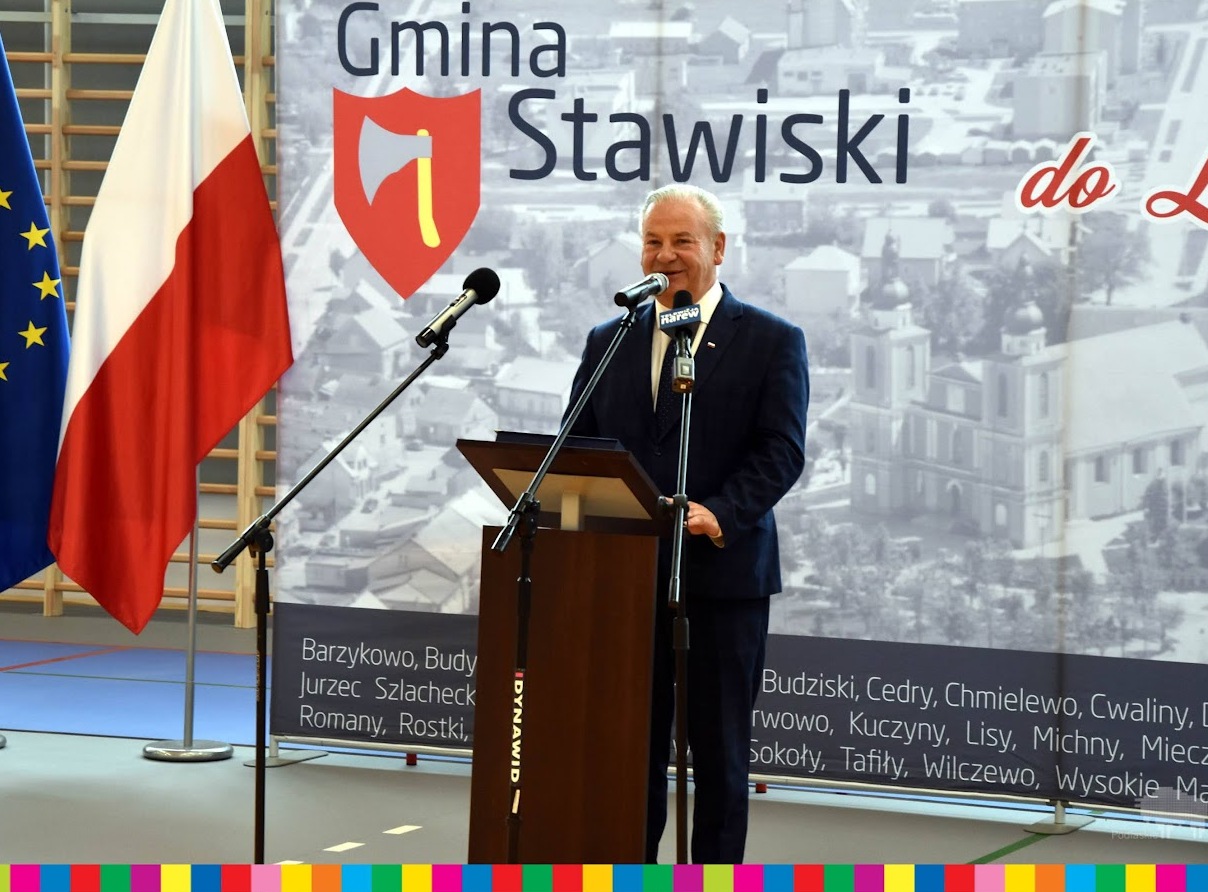 Wicemarszałek Marek Olbryś przemawia na tle grafiki z napisem Gmina Stawiski. Po lewej stronie widoczna jest flaga biało-czerwona