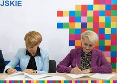 Dwie kobiety podpisują dokumenty na tle białej ścianki z pikselowym żubrem