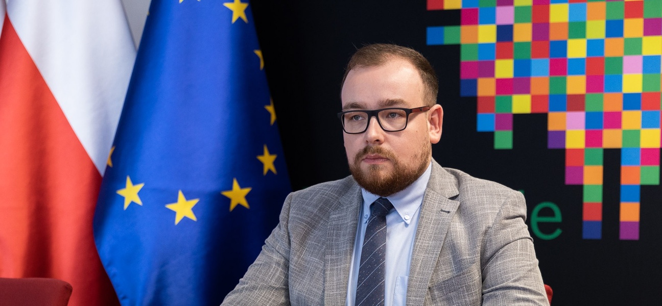 Mężczyzna w szarym garniturze. W tle czarna ścianka województwa z pikselowym żubrem, po lewej stronie flaga Unii Europejskiej i Polski