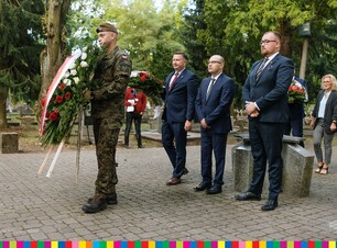 Trzech mężczyzn w garniturach stoi za żołnierzem, który trzyma wieniec