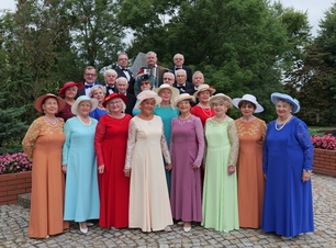 Starsze kobiety w odświętnych strojach i kapeluszach, za nimi stoją mężczyźni w garniturach